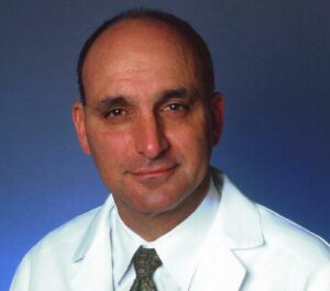 A middle-aged man with a bald head wearing a white medical coat and patterned tie, posing for a professional portrait against a blue background.