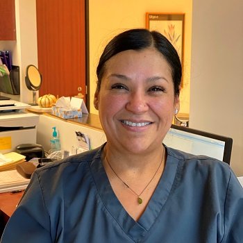A woman wearing gray scrubs smiles at the camera while seated at a desk in an office or clinic setting.