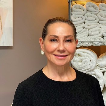 A woman with pulled-back hair and pearl earrings stands in front of shelves stacked with neatly rolled white towels.
