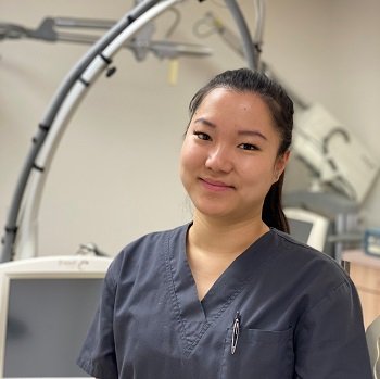 Woman in gray medical scrubs stands in a clinical setting with medical equipment and monitors in the background.