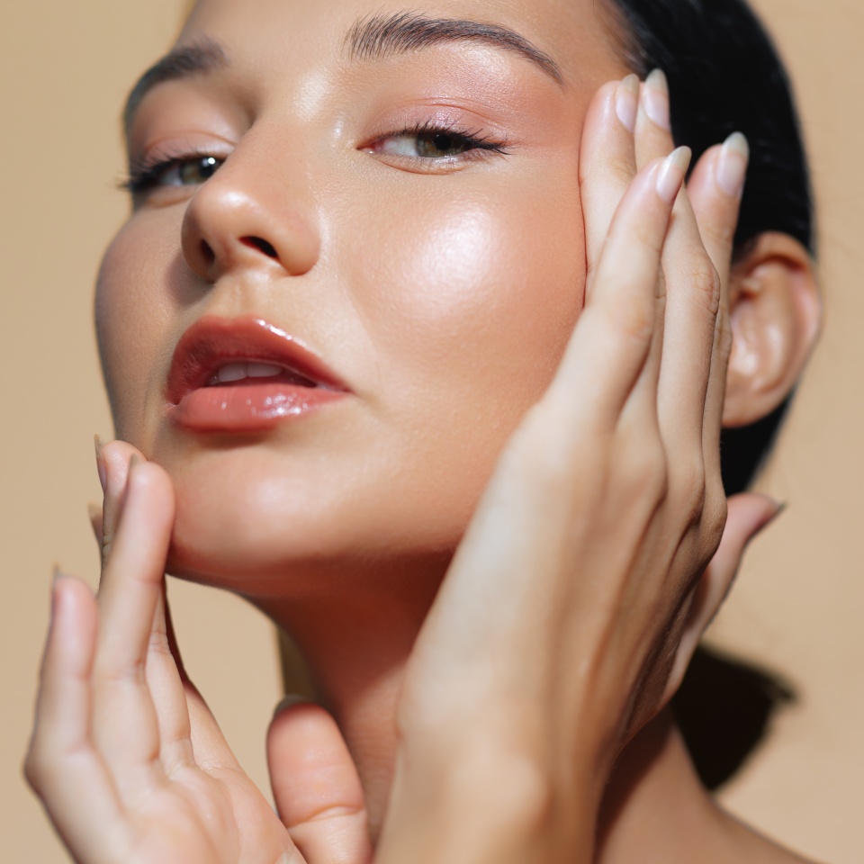 Stunning close-up of a Latina woman with glowing skin and natural makeup, gracefully caressing her face with both hands against a warm beige background. Ideal for skincare ads, beauty editorials, and