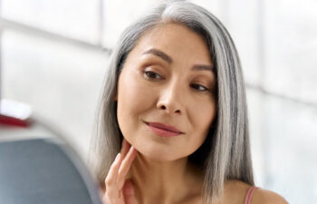 A woman with long gray hair looks at her reflection in a mirror, gently touching her face, in a softly lit room.