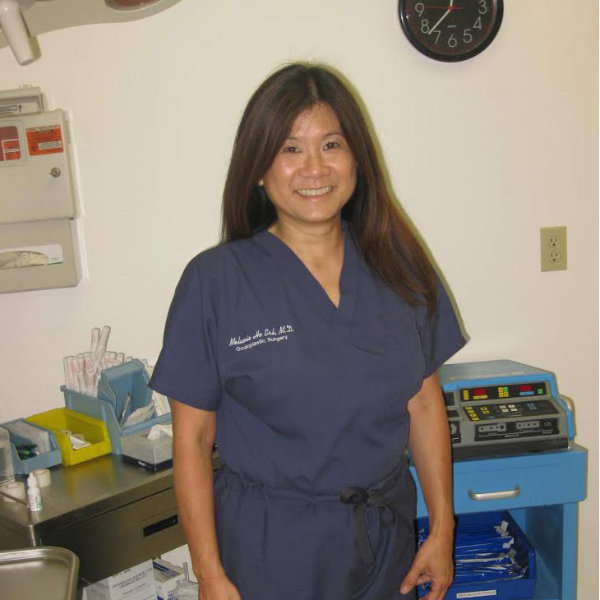 A woman in medical scrubs stands in a clinic room with medical equipment, supplies, and a clock on the wall behind her.
