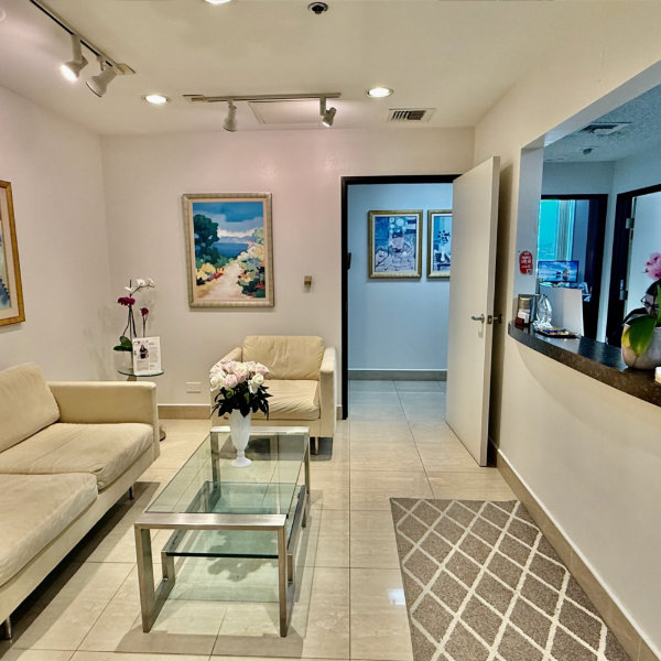A clean, well-lit waiting room with beige sofas, a glass coffee table, flower arrangements, paintings on the walls, and an open reception window.