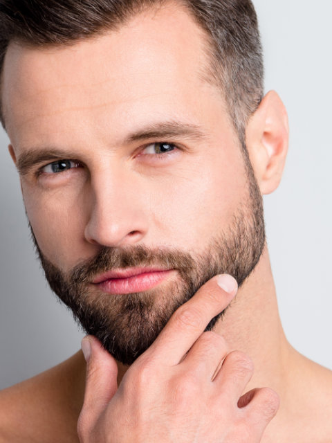 A close-up of a man with light skin, short brown hair, and a trimmed beard touching his chin, looking directly at the camera with a neutral expression.