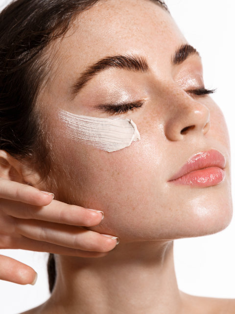 A person with closed eyes applies a white cream to their cheek, showing smooth skin, defined eyebrows, and glossy lips against a white background.