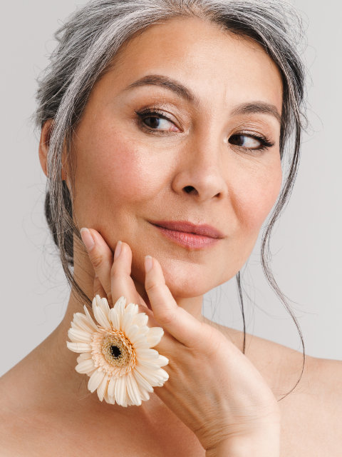 Woman with gray hair holds a large pale flower near her face, gazing to the side with a slight smile against a neutral background.
