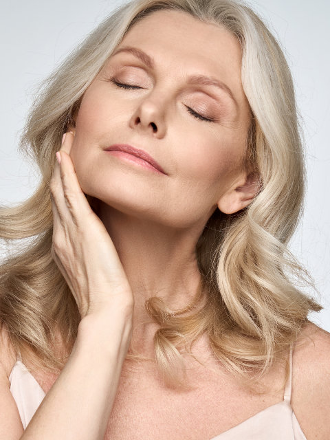 Older woman with light blonde hair touches her face gently with her eyes closed, posing against a plain background.