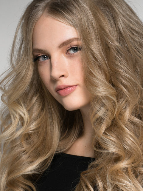 A young woman with long, wavy blonde hair and blue eyes looks at the camera. She is wearing a black top and has a neutral expression.