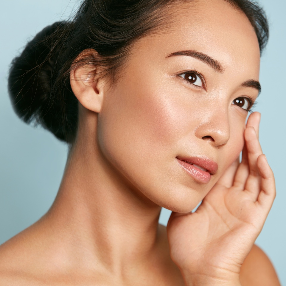 Skin care. Woman with beauty face and healthy facial skin portrait. Beautiful asian girl model with natural makeup touching glowing hydrated skin on blue background closeup. High quality image