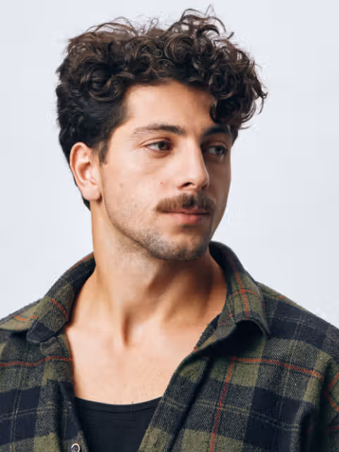 Man with curly hair and a mustache wearing a green and black plaid shirt over a black t-shirt, looking to the side against a light gray background.