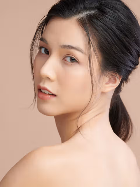 Woman with dark hair in a low ponytail looking over her shoulder, against a plain beige background, with bare shoulders visible.